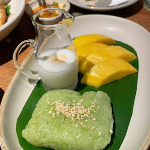 The image shows a plated dessert consisting of green sticky rice topped with sesame seeds and served on a banana leaf alongside mango slices in the ba