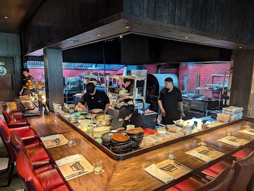 The image depicts a busy restaurant kitchen where chefs are preparing food at the counter for customers seated on red chairs in front of them.