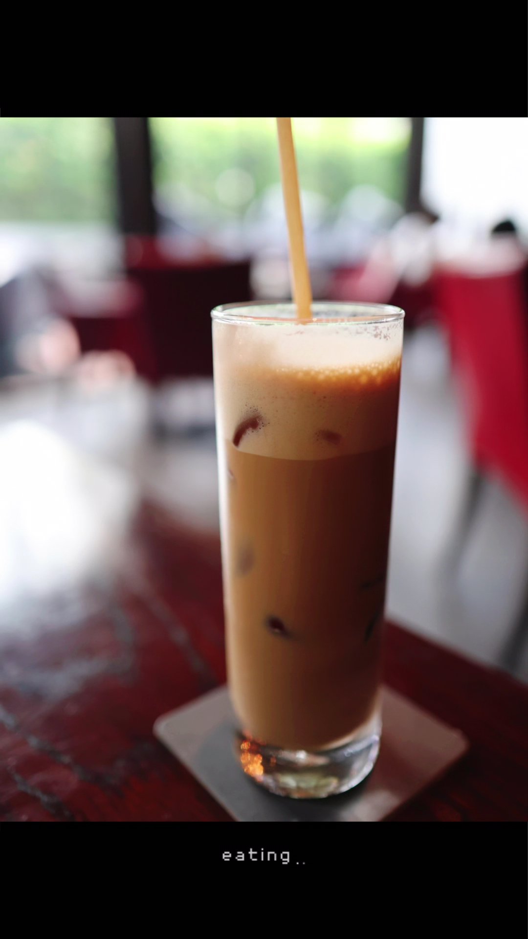 A close-up shot of a glass filled with an iced coffee beverage on a wooden table in what appears to be a cafe setting.