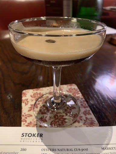 A close-up of a cocktail served in a classic coupe glass on top of a coaster with floral patterns at Stoker Wood Fired Grill & Bar, as indicated by th