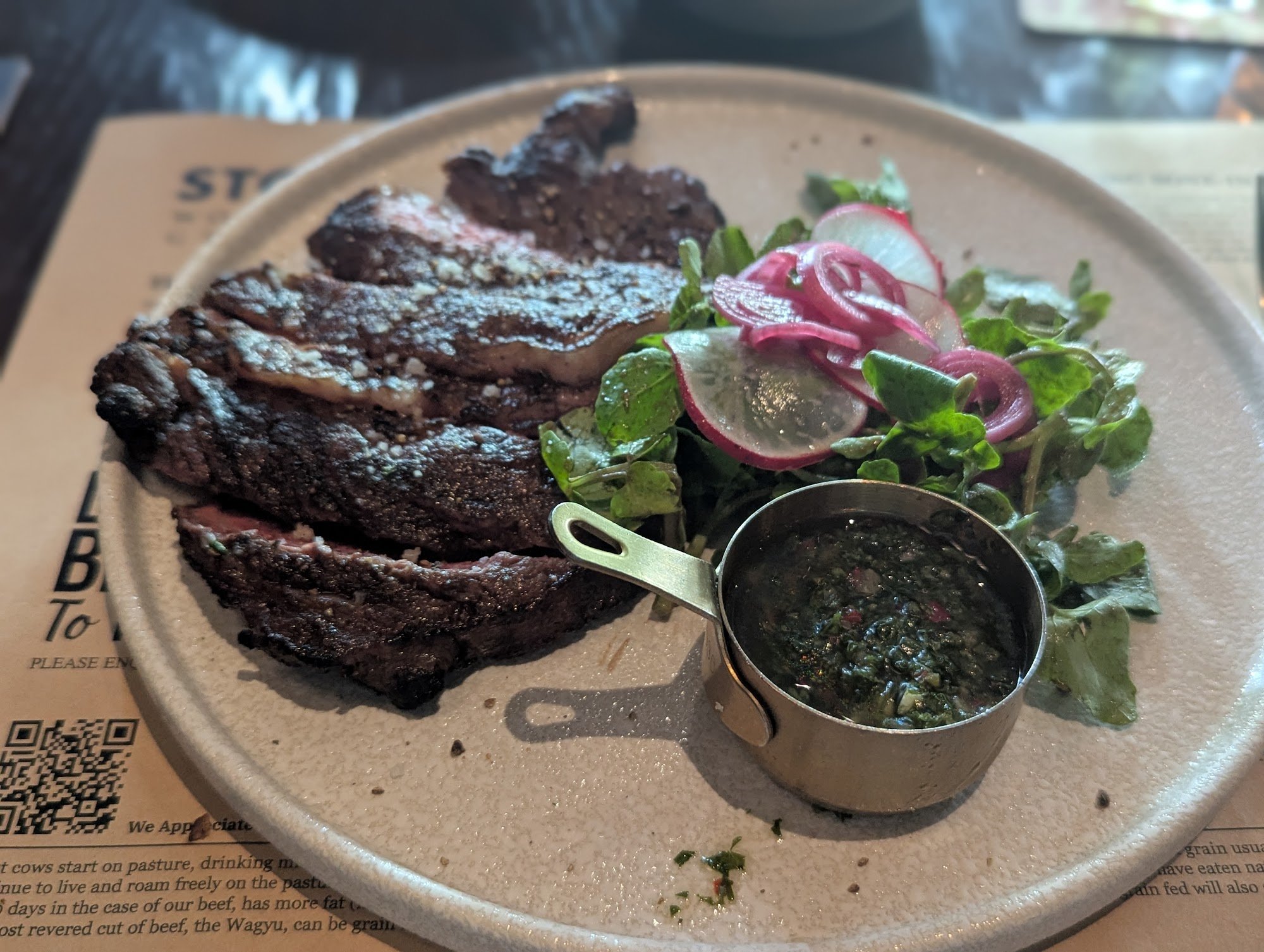 A plated dish featuring a medium-rare steak accompanied by a side salad of greens and radishes with red onion slices, served alongside a small cup con