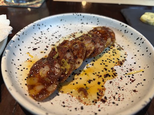 A plated dish featuring a single piece of what appears to be grilled squid or octopus with charred edges and visible spices, served on a white plate s