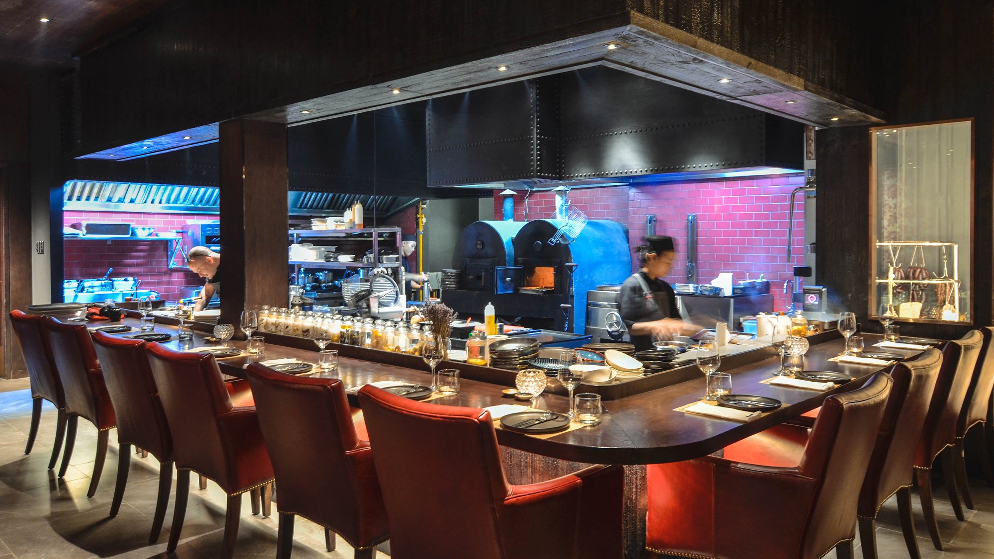 The image depicts an upscale restaurant interior featuring a long communal dining table surrounded by red leather chairs with gold accents. The bar ar