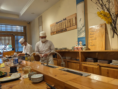 The image depicts an interior view of a Japanese-style restaurant or bar where two chefs in white uniforms and hats are working at the counter, surrou