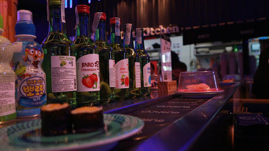 A bar counter in a restaurant/venue with various bottles (including 'TINRO STRAWBERRY' and a Japanese-style drink bottle), a plate with two dark-color
