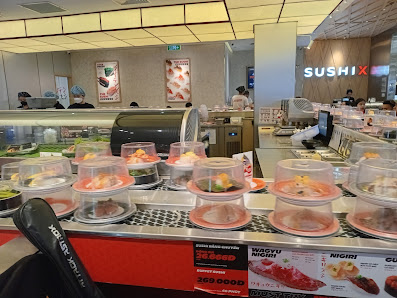 The image shows an interior view of a sushi restaurant named 'SUSHI X'. The counter displays various dishes in clear containers, with labels indicatin