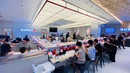 The image shows an interior view of a modern sushi restaurant named 'SUSHIX'. Patrons are seated at conveyor belt-style tables, with the kitchen area