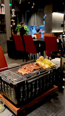 The image shows a modern restaurant interior featuring dark tones and contemporary decor. A grill with meat is in the foreground on an ornate tray, su