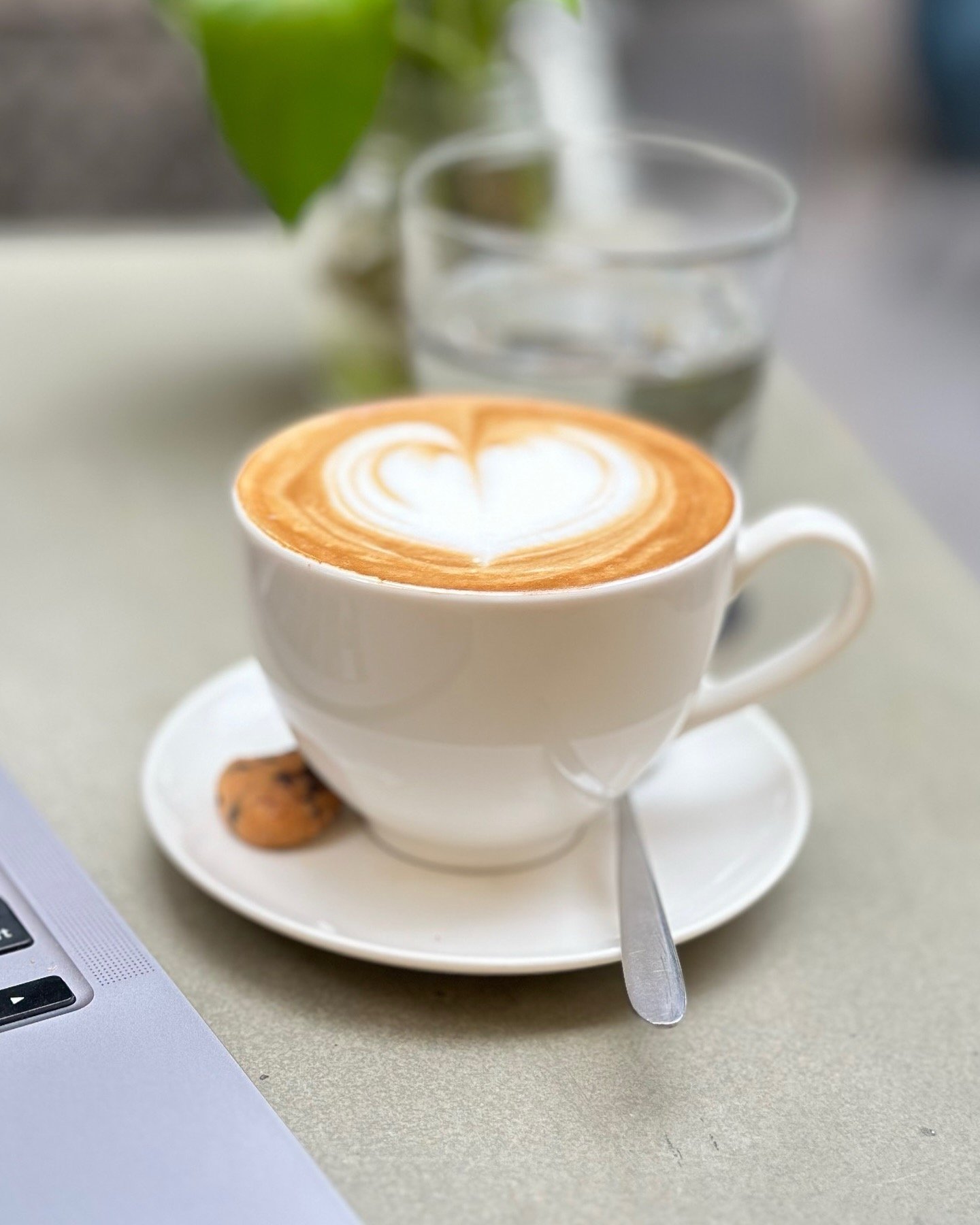 A close-up shot of a latte art coffee in a white cup on a saucer with a heart design on the foam, placed beside a laptop and a cookie on a table.