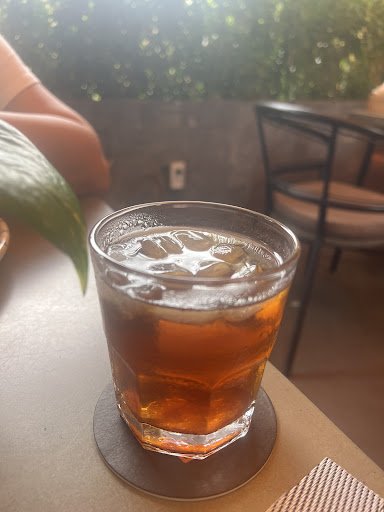 A close-up of a glass containing an amber-colored beverage on ice, possibly tea or cola, placed on a coaster at what appears to be an outdoor seating 