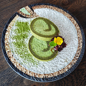 A plated dessert featuring a green-colored cake or pastry sliced into two rings on top of white rice flour dusted with matcha powder, accompanied by s