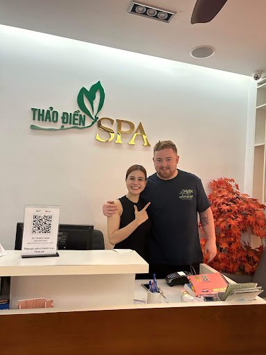 The image shows two individuals standing in front of a reception desk at Thao Dien Spa. The person on the left appears to be female, wearing black att
