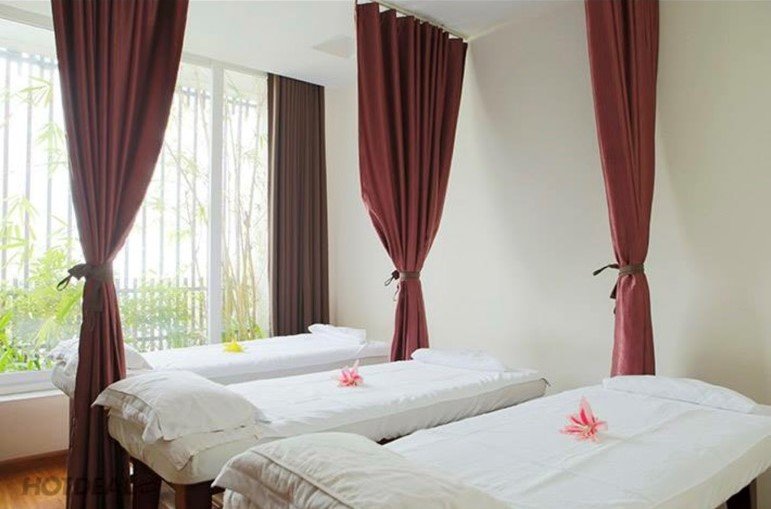 The image shows a serene and well-lit massage room in Thao Dien, Saigon with three white massage beds covered by crisp linens. Each bed is adorned wit