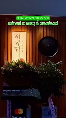 The image shows an interior view of a restaurant named 'Minari K BBQ & Seafood' with wooden paneling on the wall, potted plants, and a neon sign above