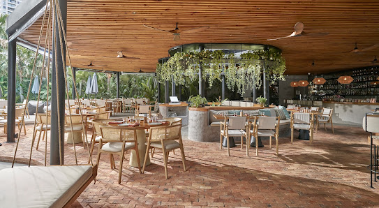 A spacious indoor-outdoor dining venue with wooden flooring and ceiling beams, featuring multiple tables with chairs, a central bar area surrounded by