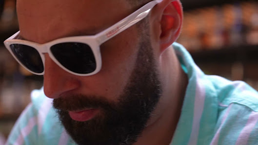 A close-up of a man wearing white-framed sunglasses and a light blue and white striped shirt, with a blurred background suggesting an indoor venue (li