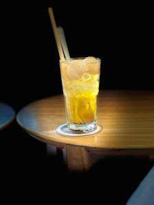 A glass of yellow-colored beverage garnished with lemon slices and a wooden straw on top is placed on a coaster atop a round table in dim lighting.