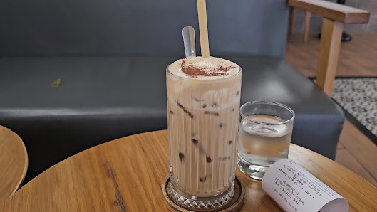 A tall glass of a layered beverage topped with what appears to be cinnamon or cocoa powder and two wooden straws is placed on a round table, accompani
