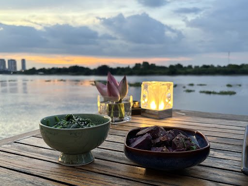 A wooden deck table by a lake at sunset in Thao Dien, Saigon, featuring a green bowl of leafy greens, a dark bowl of purple-hued food (possibly grille