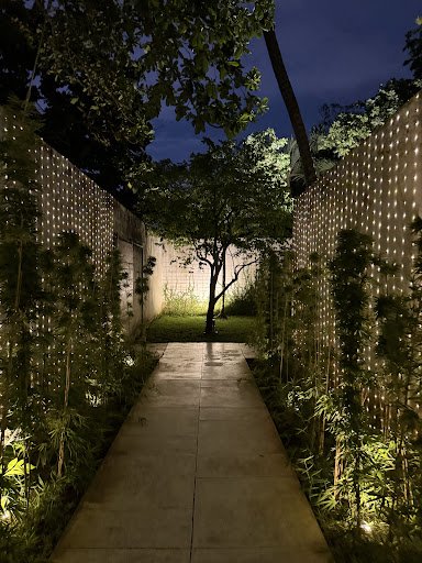 A nighttime exterior view of a serene pathway flanked by illuminated lattice walls with embedded lights, surrounded by lush greenery and bamboo plants