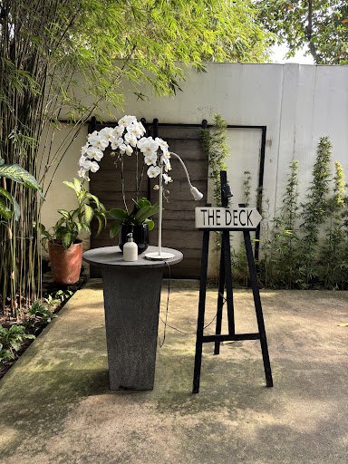 Outdoor patio area featuring a concrete table with a white orchid arrangement, a modern desk lamp, and a small container. A black tripod holds a sign