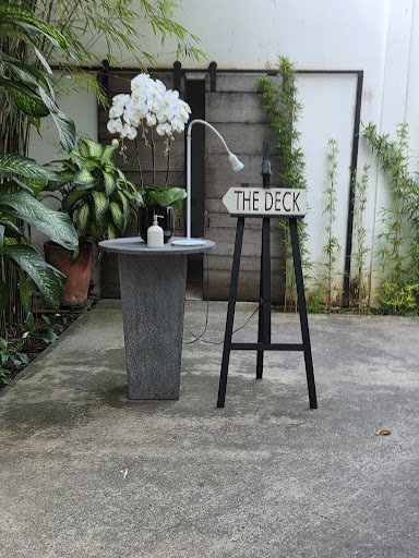 Outdoor patio area with a concrete surface, featuring a small round table with a lamp, a potted plant with white flowers, and a sign on a stand readin