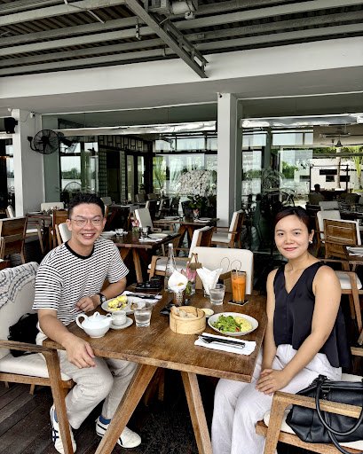 Two people seated at a wooden table in an open-air restaurant. The table features a teapot, bowls of food (including a salad and possibly rice), a bam