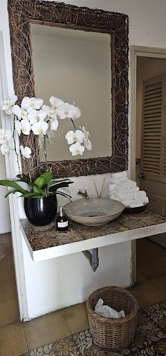 A bathroom interior featuring a large mirror with an ornate frame, a stone sink on a countertop, a black pot with white orchids, a small bottle, folde