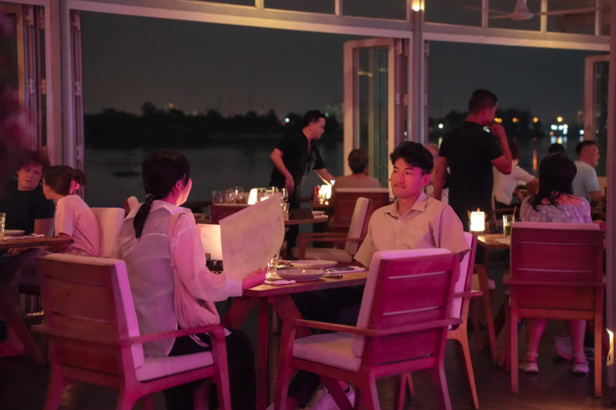 The image depicts an evening scene at an outdoor restaurant in Thao Dien, Saigon. Patrons are seated at wooden tables with cushioned chairs, engaging 