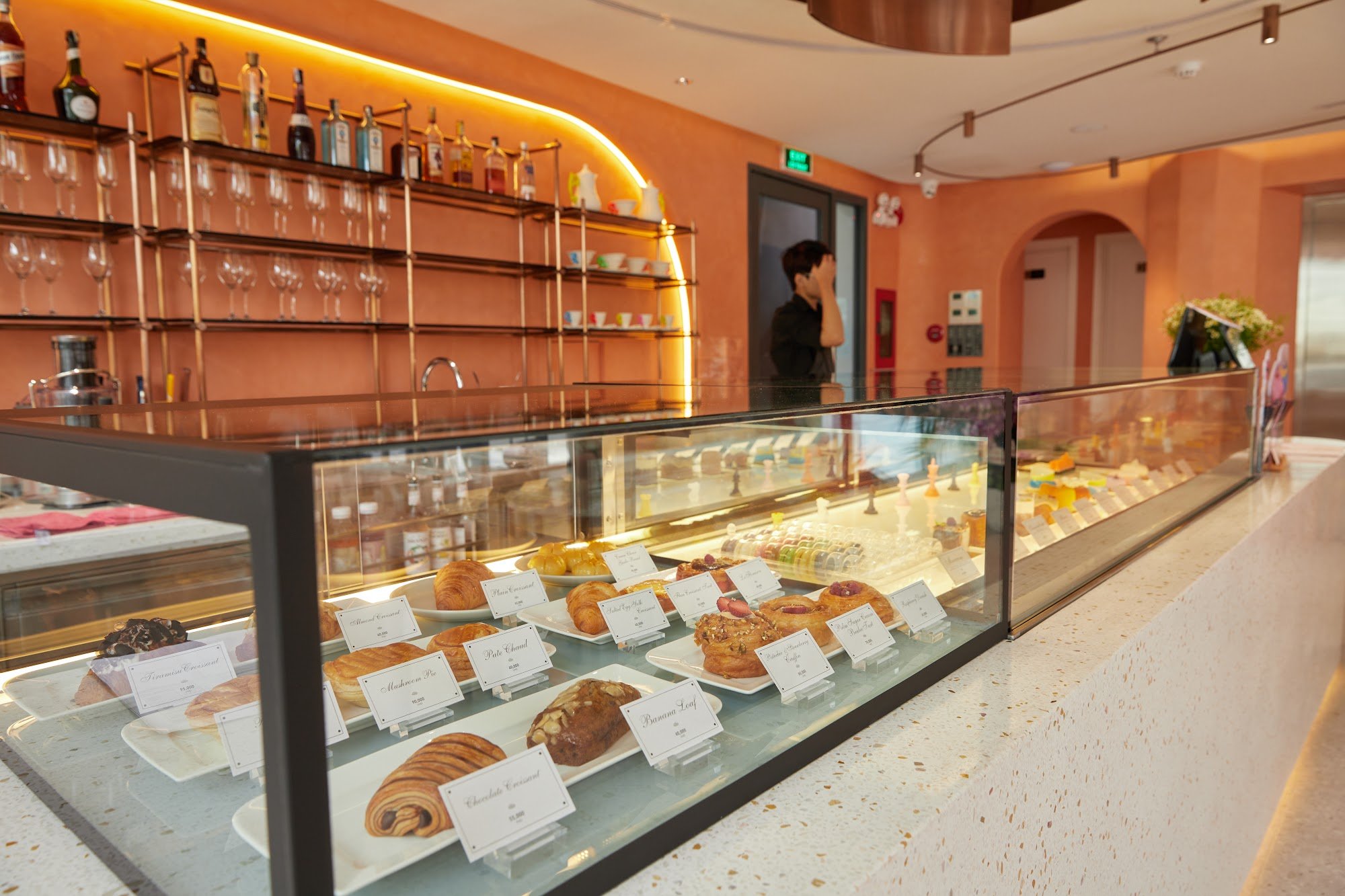 The image shows a well-lit interior of what appears to be a modern café or bakery with an emphasis on pastries and desserts displayed in a glass case