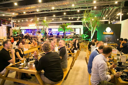 The image depicts an indoor dining area bustling with patrons seated at wooden tables enjoying meals and drinks in a lively atmosphere.