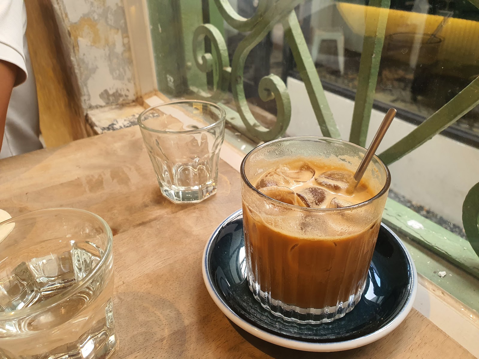 A glass of cold coffee on a saucer is placed next to an empty clear glass and another filled with water containing ice cubes, all resting on a wooden 