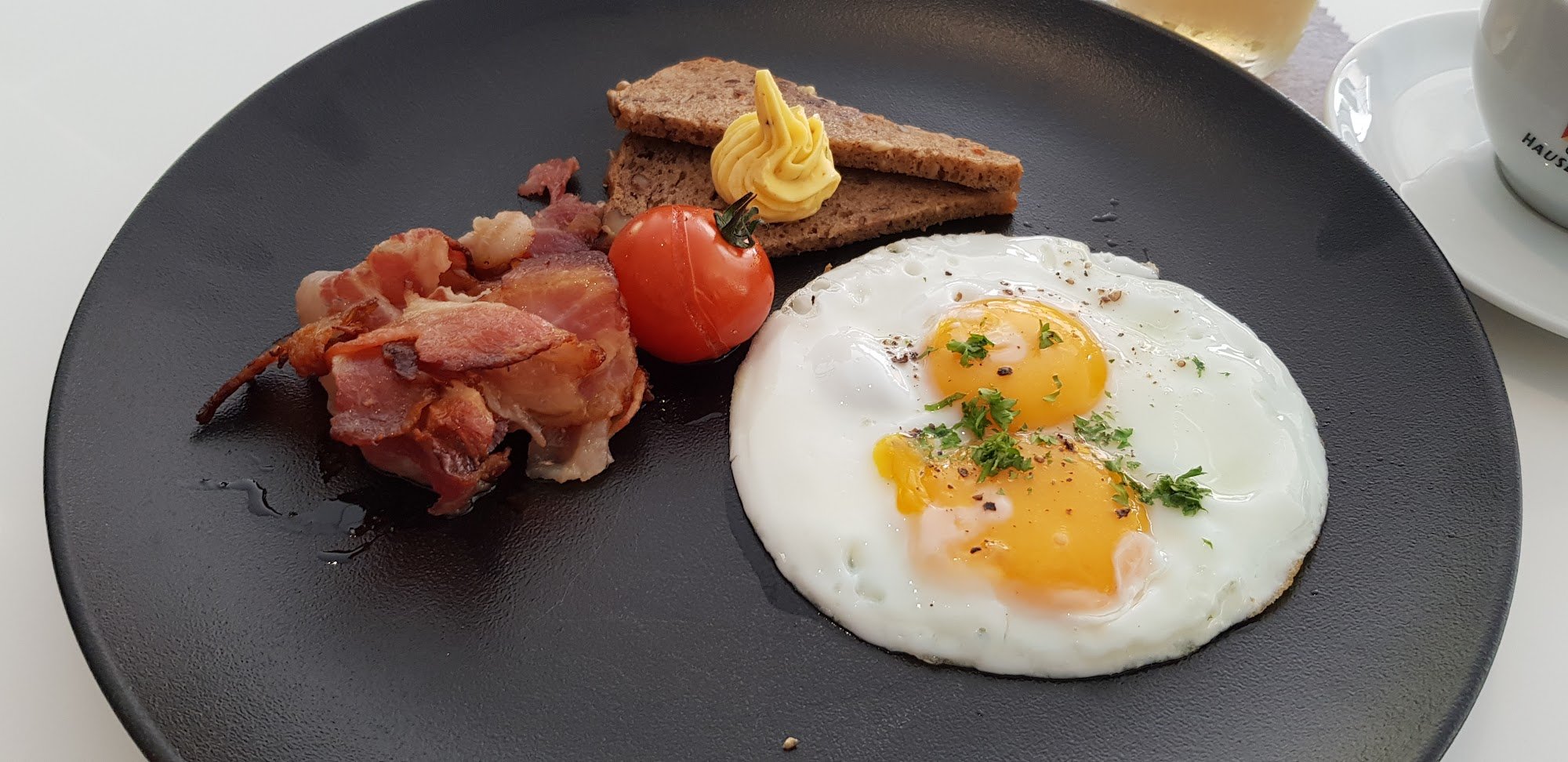 A plated breakfast meal consisting of a fried egg sunny side up with visible herbs and spices on top, accompanied by bacon strips, a slice of tomato, 