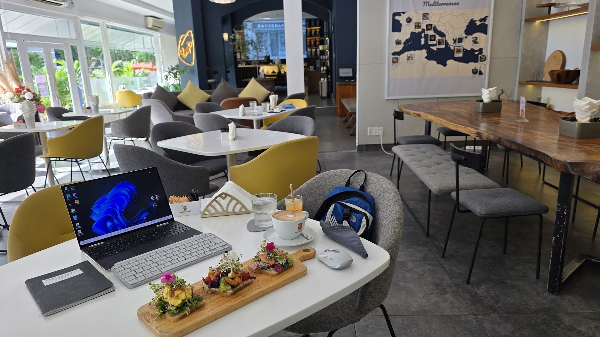The image depicts a modern café interior in Thao Dien, Saigon. It features a mix of white and yellow chairs, a long wooden communal table with gray cu