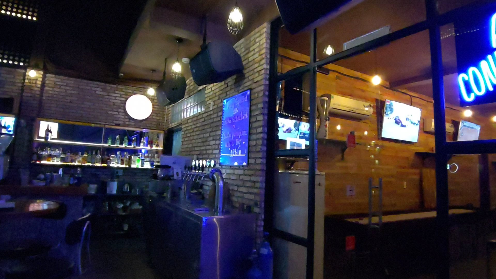 The image depicts an interior view of a bar or pub setting at night time. The establishment has exposed brick walls and modern decor, with various scr