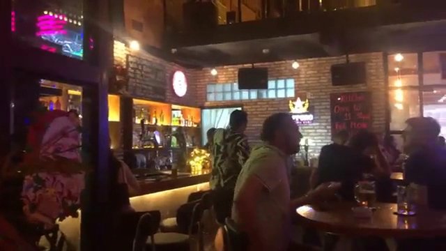 The image depicts an indoor setting of a bar or pub in Thao Dien, Saigon. The interior is dimly lit with warm ambient lighting and features exposed br