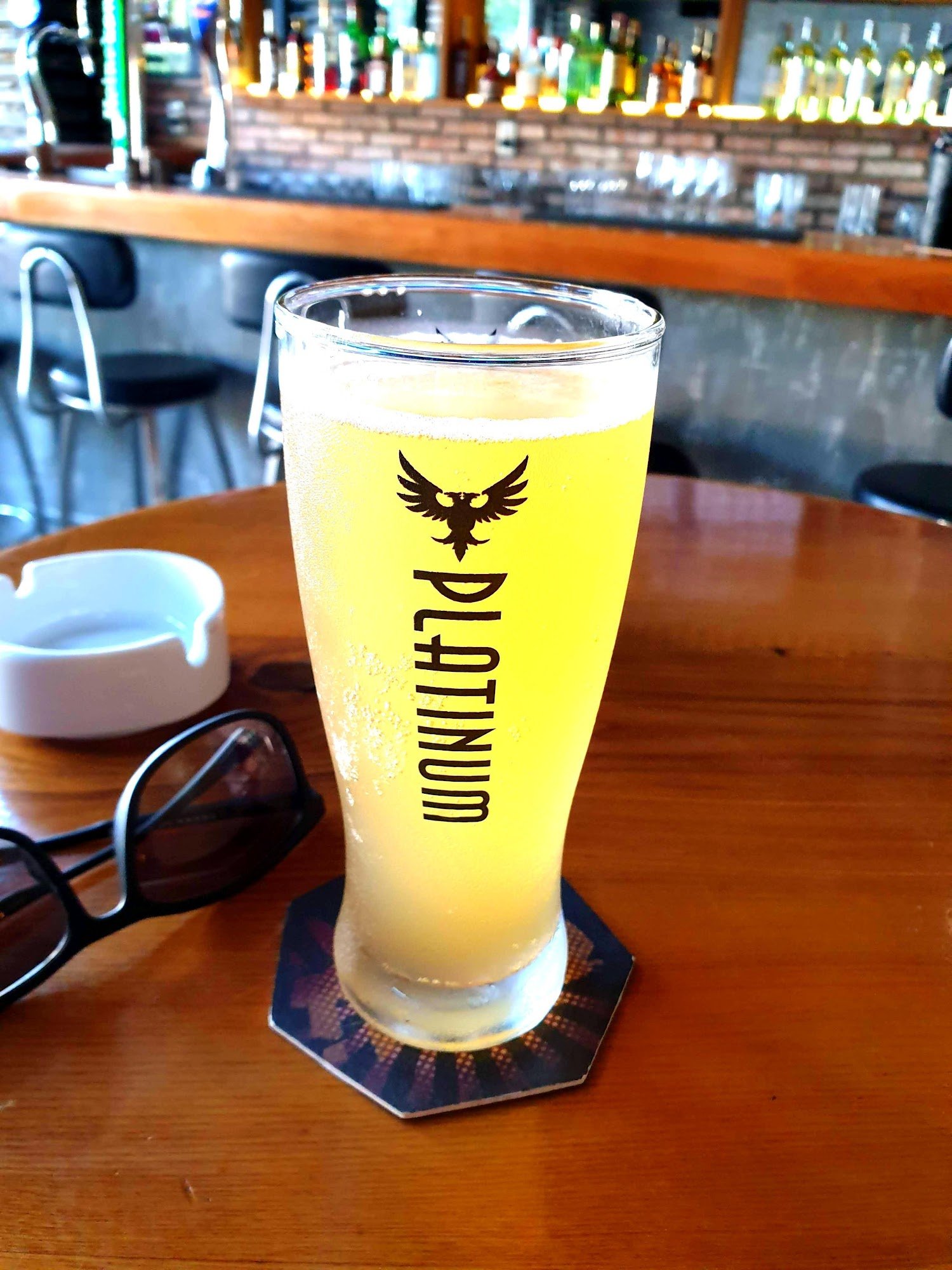 A close-up of a pint glass filled with golden beer on a coaster at an indoor bar setting.