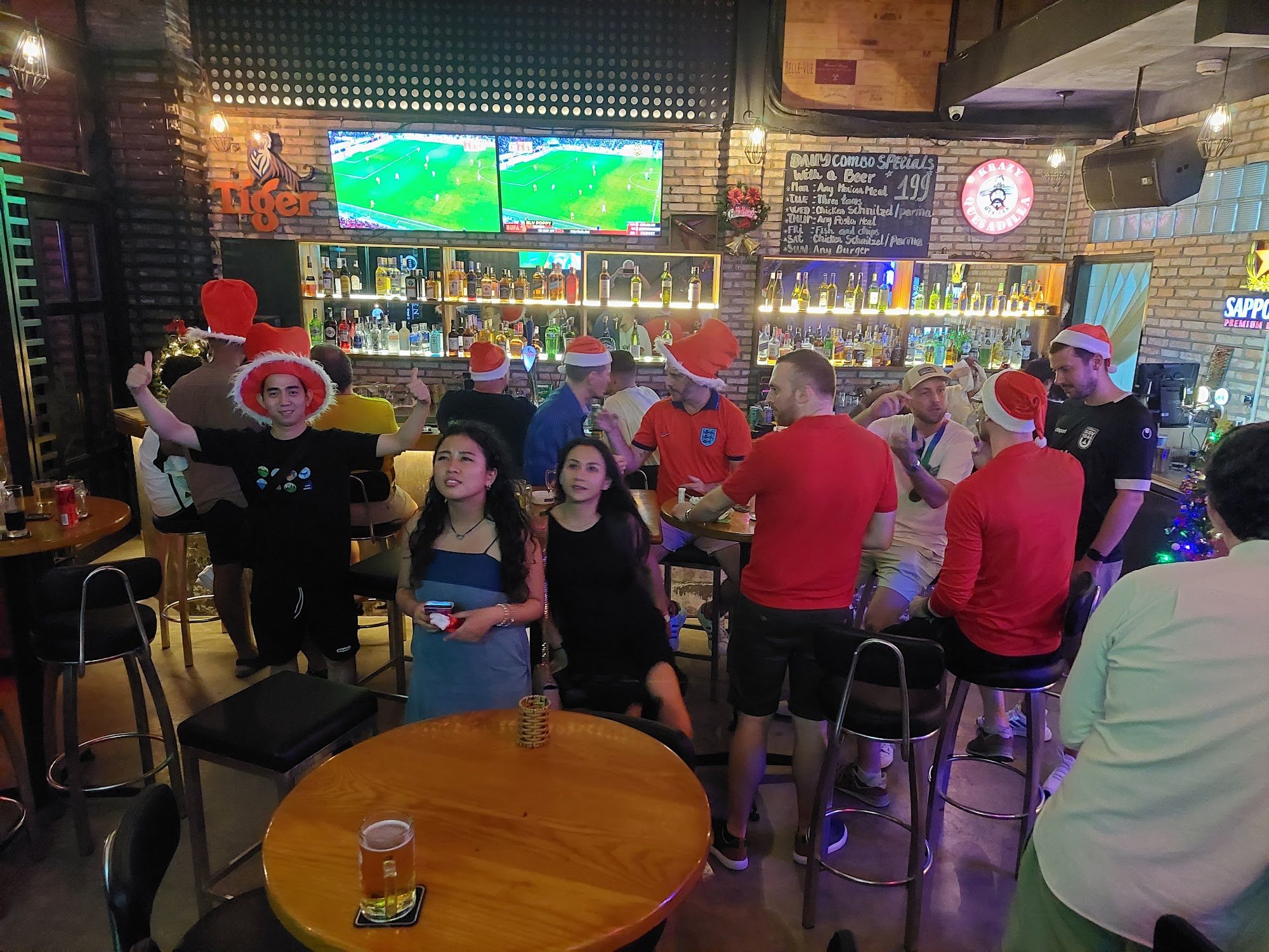 The image depicts a lively bar scene inside Thao Dien, Saigon. Several patrons are gathered around tables and stools, engaging in conversation while w