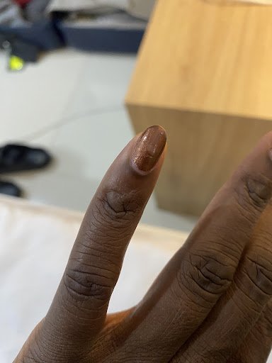 A close-up photograph of a person's hand showing their fingers and nails in an indoor setting with visible furniture pieces like a wooden box or table