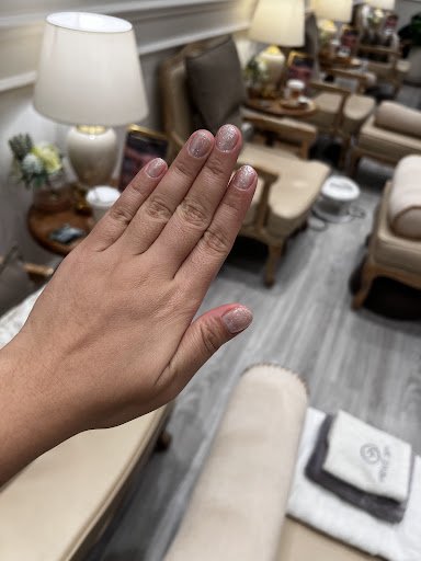 A close-up of a person's hand displaying freshly manicured nails in what appears to be an upscale nail salon or spa, with well-lit and furnished waiti