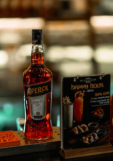 A close-up of a bar setting featuring a bottle of Aperol (a red-orange Italian aperitif) and a menu board advertising 'brunch hour' with 50% off on ce
