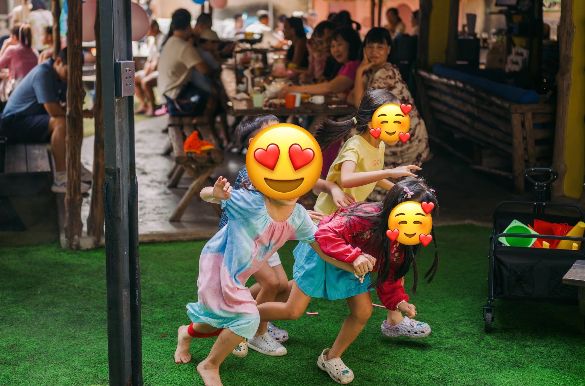 Four children are playing on artificial grass in an outdoor area of a venue. They have emoji overlays on their faces. In the background, there are adu