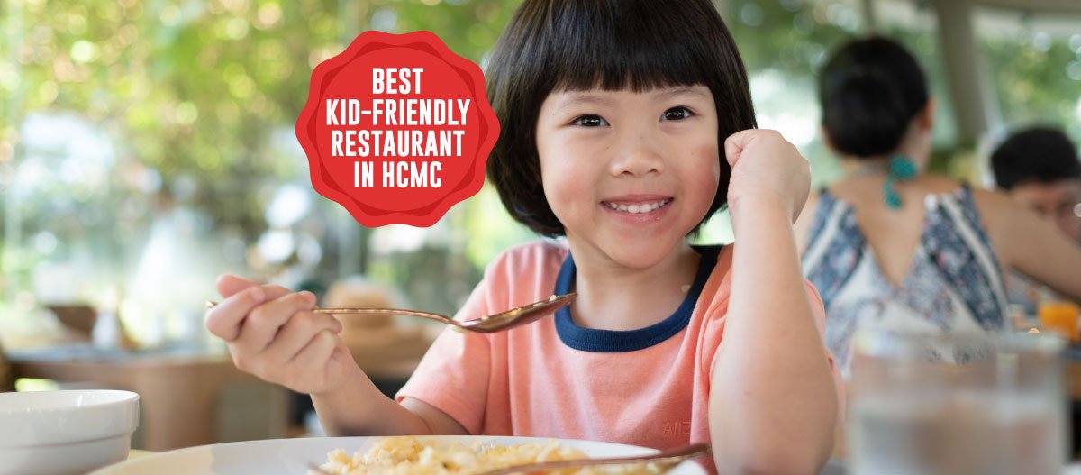 A child in a pink shirt is eating at an outdoor restaurant table, holding a spoon. The background shows other diners and greenery, with a red badge st