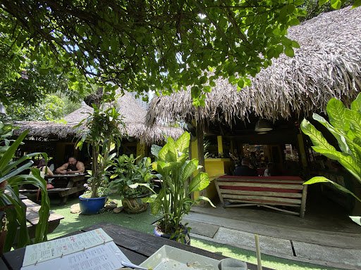 The image depicts an outdoor seating area of a tropical-themed establishment featuring thatched roofs and lush greenery. The setting appears relaxed, 