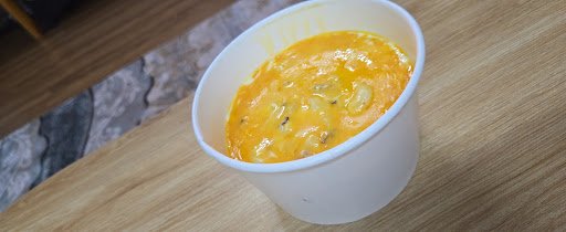 A close-up of a bowl containing an orange-colored soup or stew in a disposable white cup on a wooden surface.