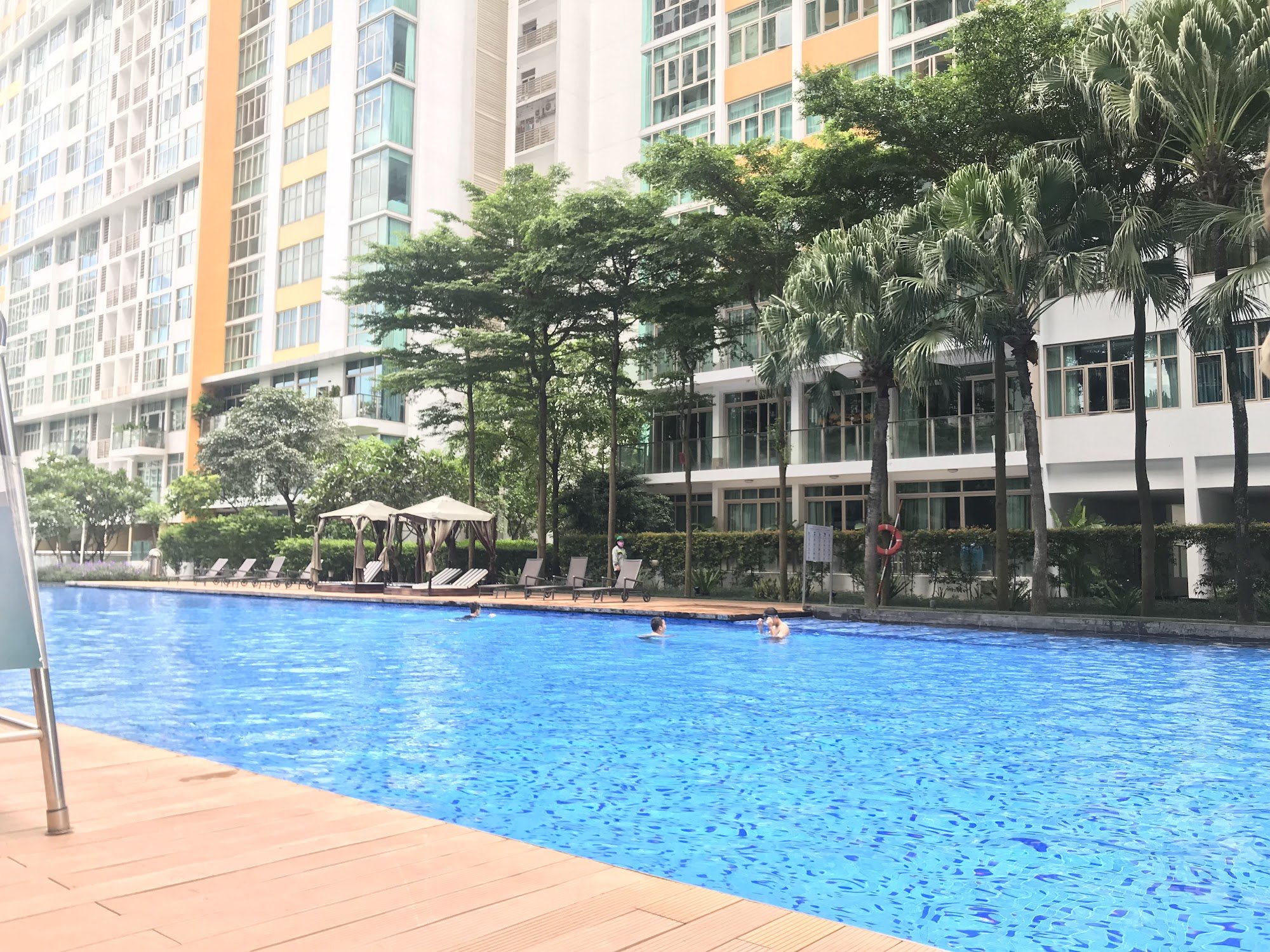 The image shows a large outdoor swimming pool surrounded by tall buildings and lush greenery in what appears to be an urban residential area.
