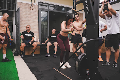 The image depicts a gym setting where several individuals are engaged in various exercises using fitness equipment such as rowing machines and free we