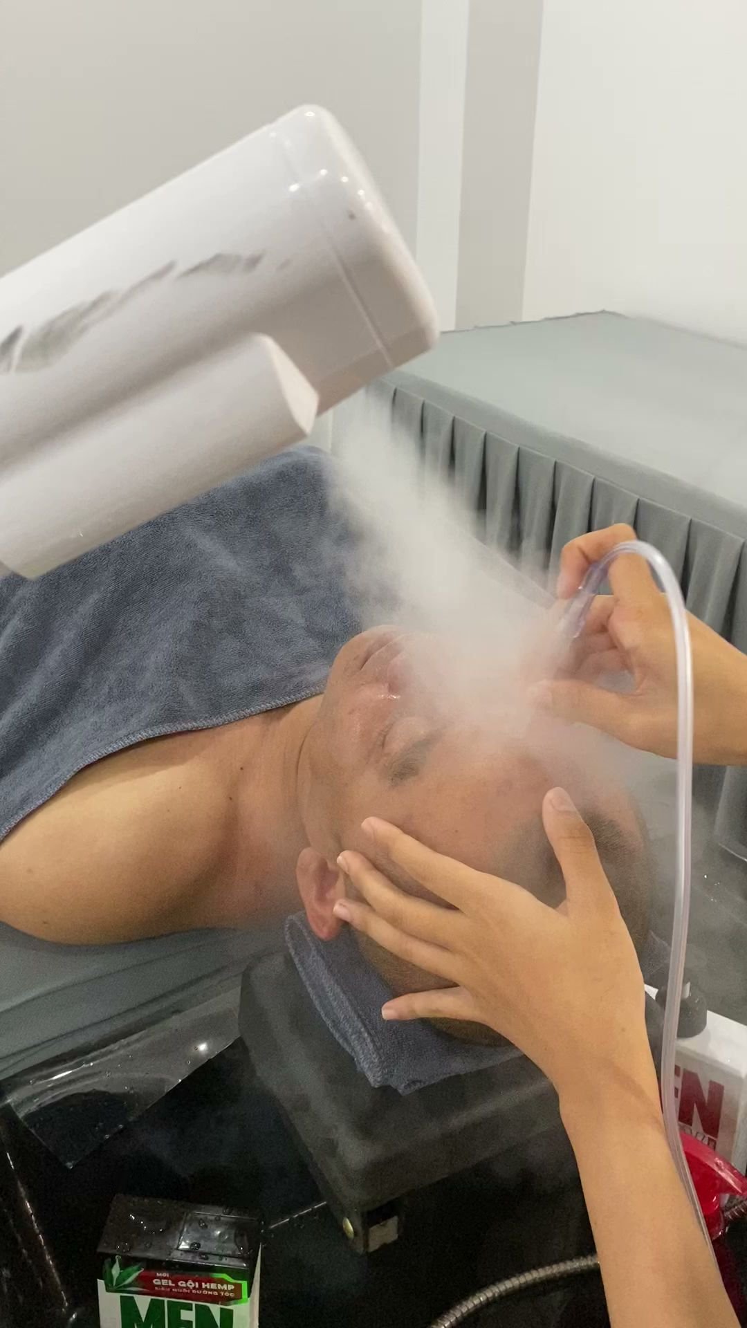 A person is undergoing a facial treatment involving the use of steam from an industrial-looking device while lying down on what appears to be a massag
