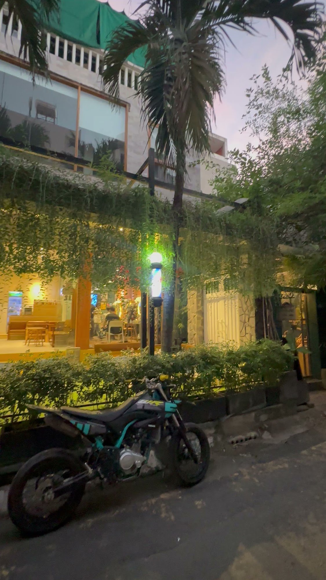 The image depicts the exterior of a venue in Thao Dien, Saigon, during twilight. A black and teal motorcycle is parked on the street in front of the e