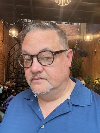 A man wearing glasses and a blue polo shirt is standing in an indoor setting that appears to be a restaurant or cafe with exposed brick walls, hanging